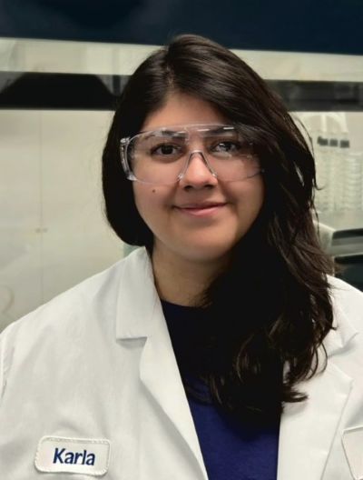 smiling woman with dark hair wearing goggles and a lab coat.in a lab coat.