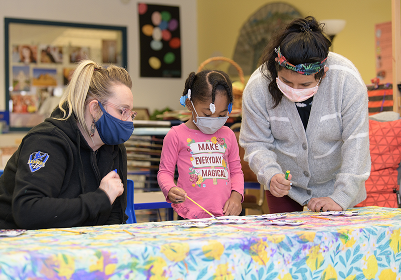 Two women helping young girl with paints